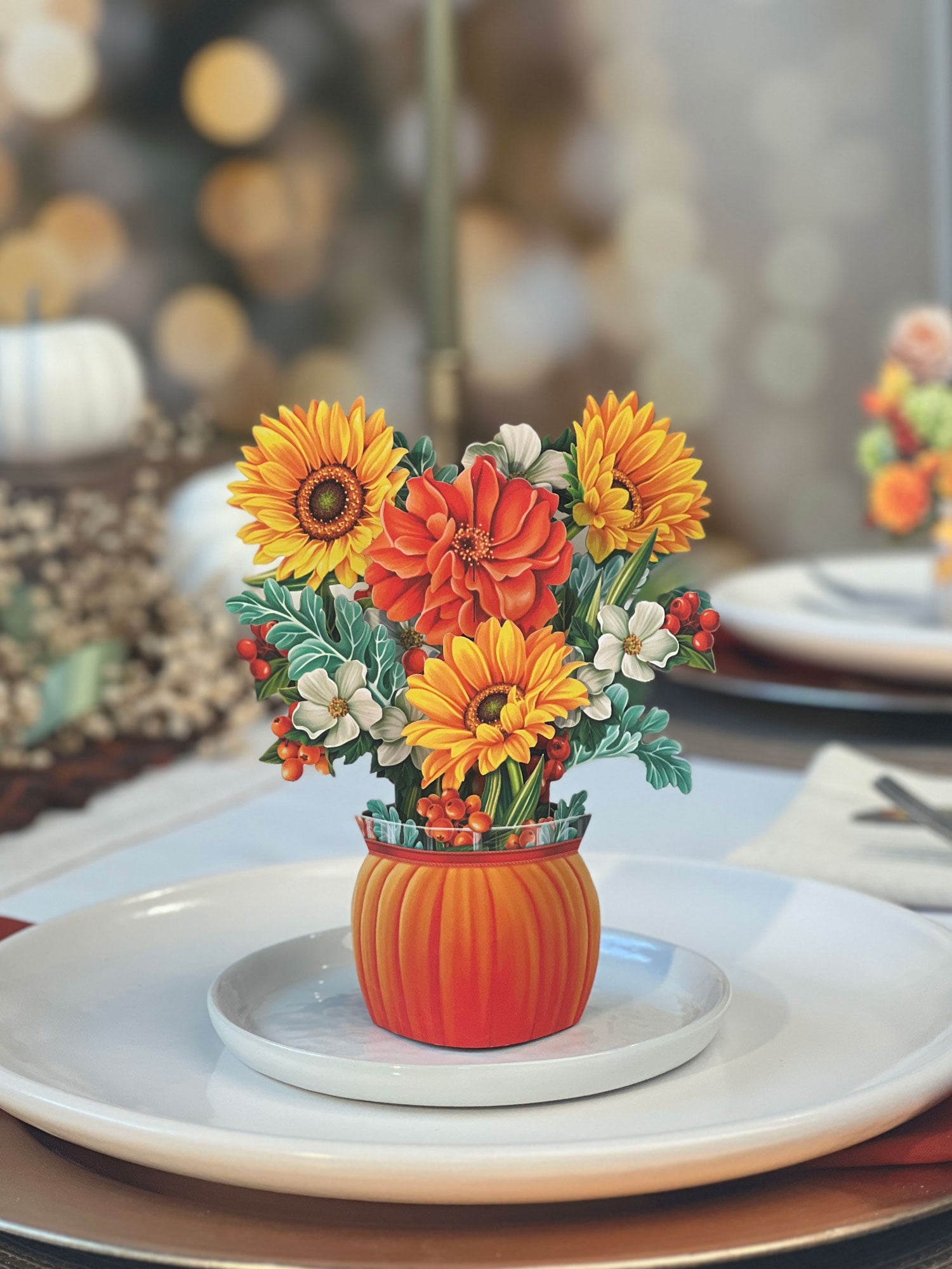 The Mini Pumpkin Harvest by Freshcut Paper is a festive autumn centerpiece showcasing an arrangement of vibrant fall flowers in shades of orange, yellow, and white, including sunflowers and dahlias. These beautiful blooms are nestled in an orange vase shaped like a pumpkin and rest on a white plate as part of a table setting with blurred lights in the background.