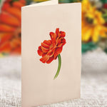 A Mini Pumpkin Harvest greeting card from Freshcut Paper stands upright on a textured surface, showcasing a simple yet vibrant illustration of fall flowers with red and orange petals and green stems. The background is a blur of bright colors, possibly representing a variety of autumn blossoms in an orange vase.