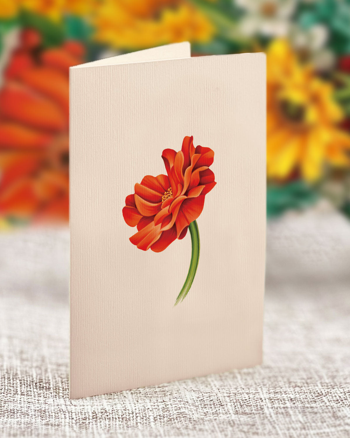 A Mini Pumpkin Harvest greeting card from Freshcut Paper stands upright on a textured surface, showcasing a simple yet vibrant illustration of fall flowers with red and orange petals and green stems. The background is a blur of bright colors, possibly representing a variety of autumn blossoms in an orange vase.