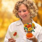 A woman with curly blonde hair smiles while reading a card. She holds Freshcut Paper's Mini Pumpkin Harvest, a small floral arrangement in an orange, pumpkin decor vase. She is wearing a white shirt and bracelets. The background is blurred with warm, golden autumn tones.