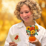 A woman with curly blonde hair smiles while reading a card. She holds Freshcut Paper's Mini Pumpkin Harvest, a small floral arrangement in an orange, pumpkin decor vase. She is wearing a white shirt and bracelets. The background is blurred with warm, golden autumn tones.