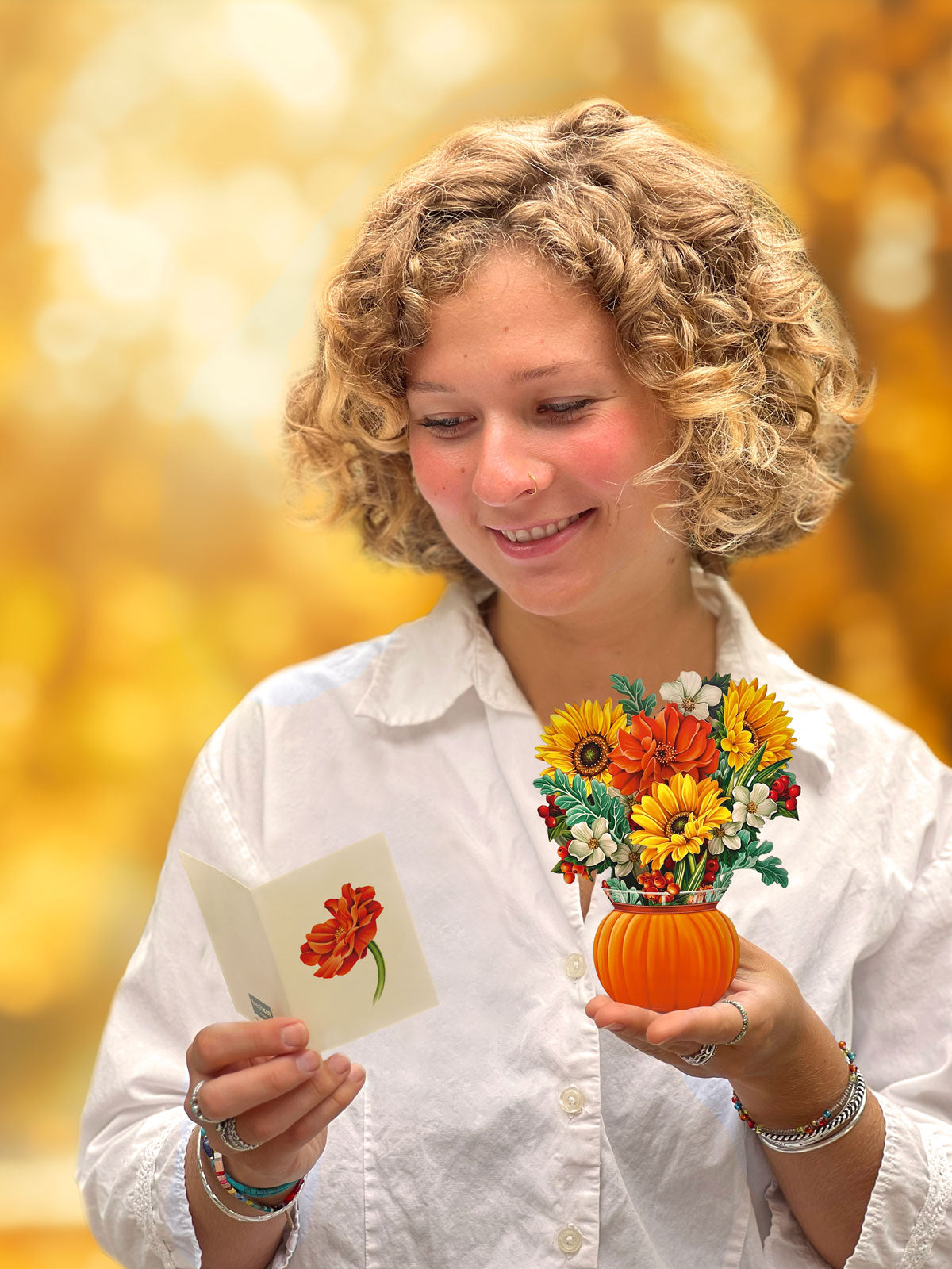 A woman with curly blonde hair smiles while reading a card. She holds Freshcut Paper's Mini Pumpkin Harvest, a small floral arrangement in an orange, pumpkin decor vase. She is wearing a white shirt and bracelets. The background is blurred with warm, golden autumn tones.