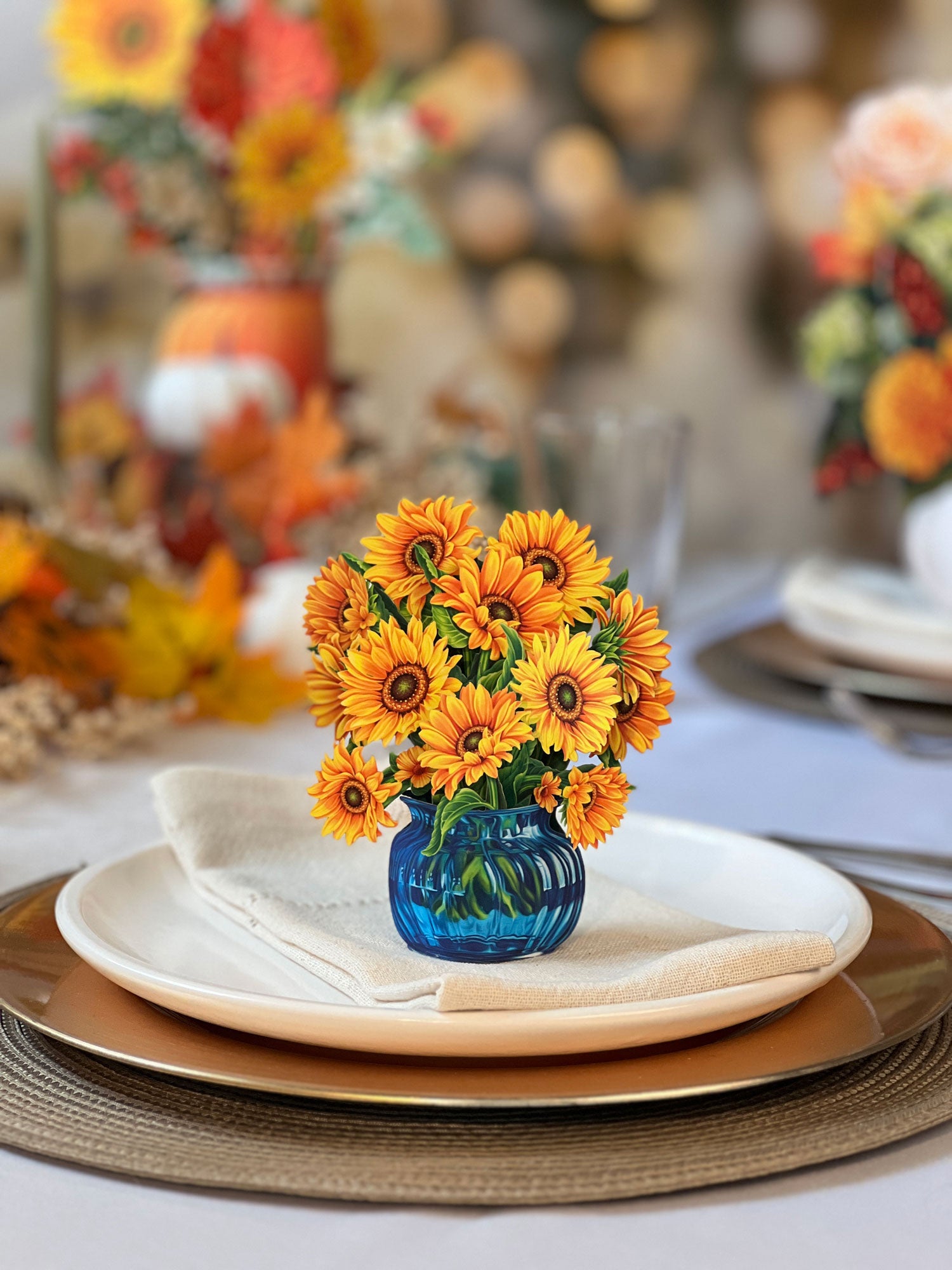 A table is set with white and brown plates, a folded cloth napkin, and a blue glass vase filled with vibrant Freshcut Paper Mini Sunflowers. In the background, there are more floral arrangements and blurred lights creating a warm, festive atmosphere.